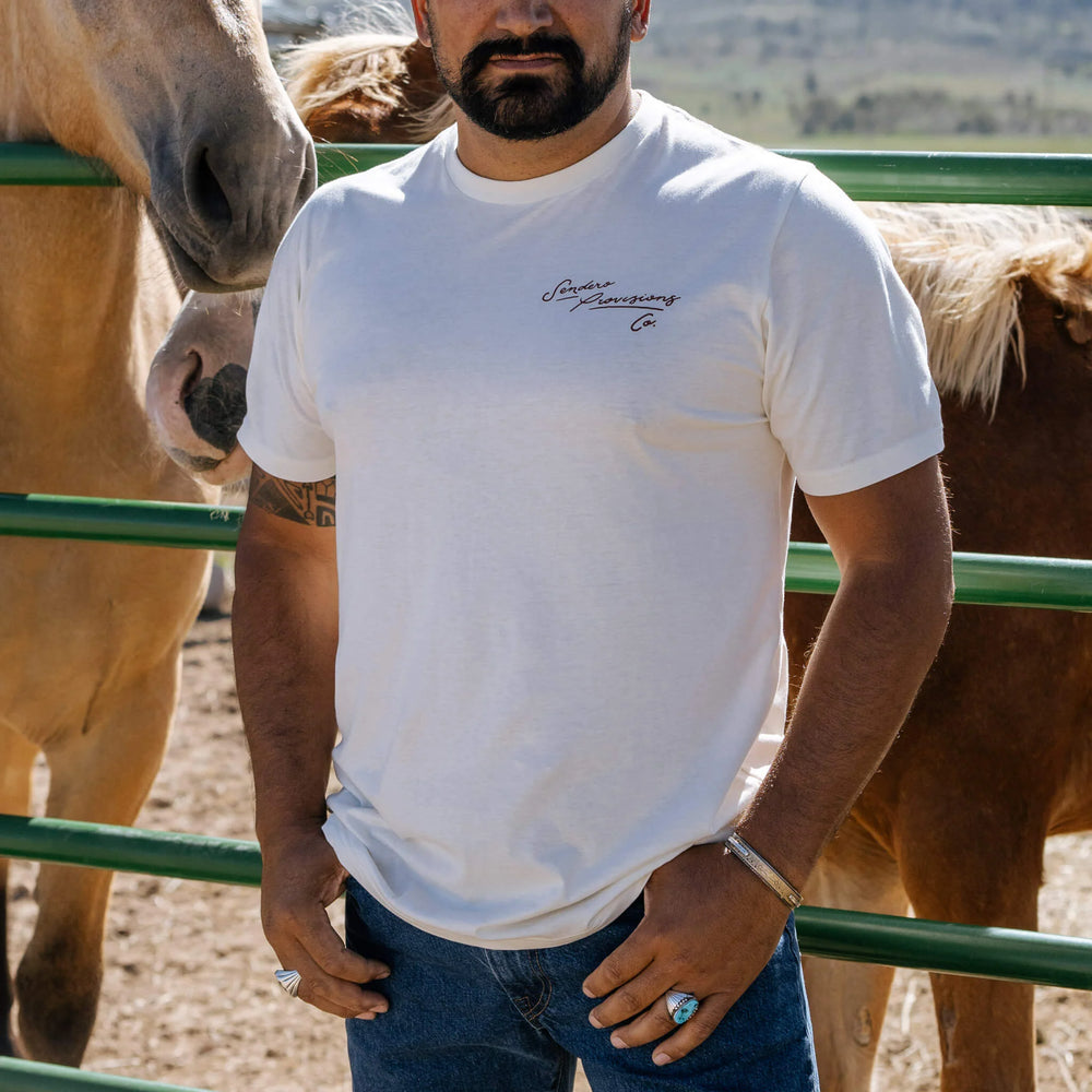 Man wearing a white t-shirt with text standing next to horses in a fenced area.
